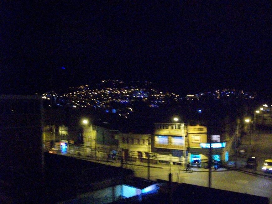 the buildings on the street are lit by streetlights, and the hills beyond are a forest of twinkling lights